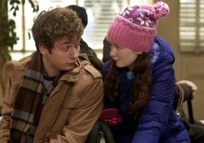 Jovem e garota em roupas de inverno. Ele de jaqueta marrom e cachecol, ela com gorro rosa e casaco azul. Ambiente interno, clima pensativo.