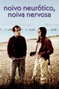 Um homem de óculos sério e uma mulher sorridente estão em uma paisagem árida,com água e construções ao fundo. Atmosfera leve.