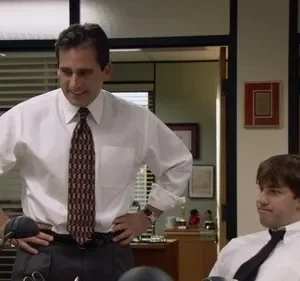 Em um escritório, um homem de pé sorri presunçosamente para outro sentado, que o observa com uma expressão divertida e cética.