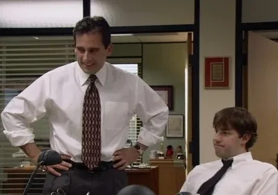 Em um escritório, um homem de pé sorri presunçosamente para outro sentado, que o observa com uma expressão divertida e cética.