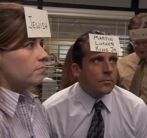 Funcionários em um escritório com etiquetas na testa, como Jewish e Martin Luther King Jr.. Olhares confusos em um ambiente de trabalho.