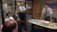 Em reunião de escritório, um homem de óculos e gravata, em pé, fala com colegas sentados. Mesa com papéis. Ambiente formal de trabalho.