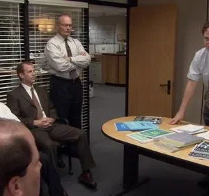 Em reunião de escritório, um homem de óculos e gravata, em pé, fala com colegas sentados. Mesa com papéis. Ambiente formal de trabalho.