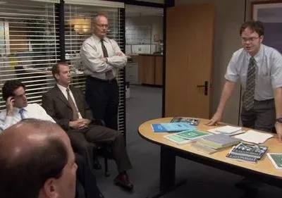 Em reunião de escritório, um homem de óculos e gravata, em pé, fala com colegas sentados. Mesa com papéis. Ambiente formal de trabalho.