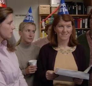 Grupo de colegas em festa de aniversário no escritório. Uma mulher sorri com papéis; outras parecem sérias, usando chapéus de festa.