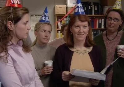 Grupo de colegas em festa de aniversário no escritório. Uma mulher sorri com papéis; outras parecem sérias, usando chapéus de festa.