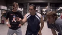 Três homens em roupas esportivas num armazém. O do centro aponta, enquanto os outros dois o observam com atenção.