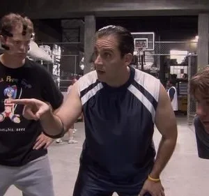 Três homens em roupas esportivas num armazém. O do centro aponta, enquanto os outros dois o observam com atenção.