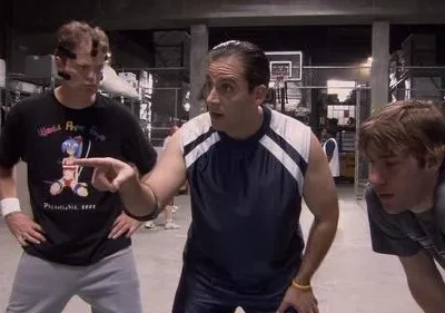 Três homens em roupas esportivas num armazém. O do centro aponta, enquanto os outros dois o observam com atenção.