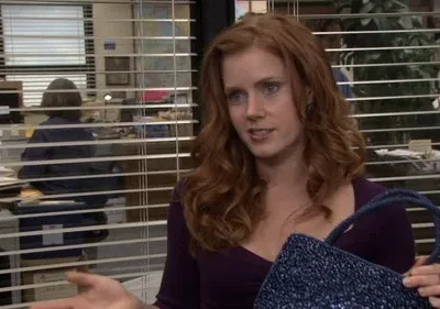 Mulher ruiva de top roxo, olhar atento, segura bolsa azul. Fundo de escritório com persianas e pessoa sentada.