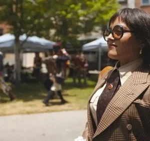 Mulher de óculos escuros e blazer xadrez marrom, em primeiro plano. Ao fundo, evento histórico com pessoas em uniformes e barracas.
