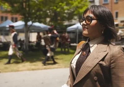 Mulher de óculos escuros e blazer xadrez marrom, em primeiro plano. Ao fundo, evento histórico com pessoas em uniformes e barracas.