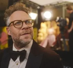 Homem de óculos e barba, de smoking, em evento de gala com luzes e câmeras ao fundo. Olhar pensativo.