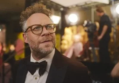 Homem de óculos e barba, de smoking, em evento de gala com luzes e câmeras ao fundo. Olhar pensativo.