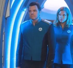 Pôster: homem e mulher em uniformes azuis de ficção científica, com semblantes sérios, num corredor futurista iluminado em azul.