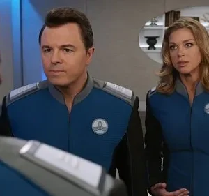Dois oficiais de uniforme azul e preto, um homem sério e uma mulher surpresa, conversam num cenário futurista.
