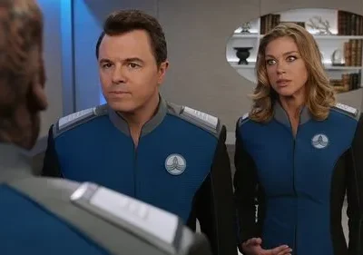 Dois oficiais de uniforme azul e preto, um homem sério e uma mulher surpresa, conversam num cenário futurista.