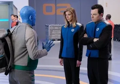 Um alienígena azul de costas gesticula para dois tripulantes com uniformes futuristas azuis, em uma base espacial clara.