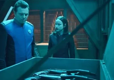 Um homem de uniforme azul e uma mulher com marcas faciais observam o conteúdo de uma caixa num ambiente futurista azul-petróleo.