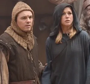 Um homem de capuz e uma mulher encapuzada observam com preocupação, ao lado de um ancião barbudo. Trajes históricos.