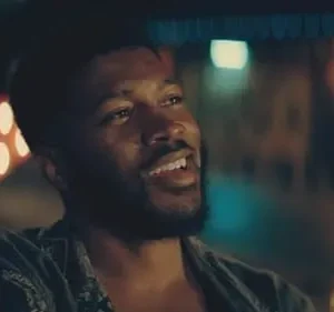 Homem negro barbudo sorrindo, olhando para o lado. Cenário noturno com luzes coloridas desfocadas ao fundo. Clima alegre.