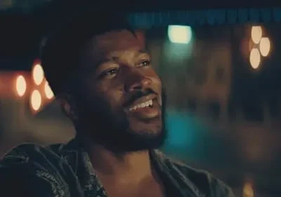 Homem negro barbudo sorrindo, olhando para o lado. Cenário noturno com luzes coloridas desfocadas ao fundo. Clima alegre.