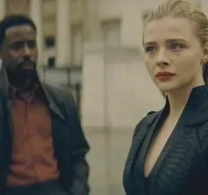 Mulher loira séria em casaco escuro, homem de casaco e camisa laranja desfocado ao fundo. Cena urbana com clima sombrio.