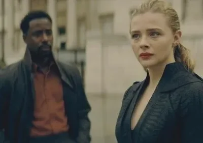 Mulher loira séria em casaco escuro, homem de casaco e camisa laranja desfocado ao fundo. Cena urbana com clima sombrio.