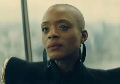 Mulher negra careca, olhar penetrante, com brincos grandes e roupa escura estruturada. Fundo urbano embaçado sugere vista alta.