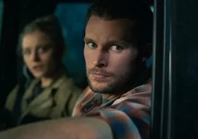 Homem barbudo de olhar intenso, camisa xadrez. Mulher séria e desfocada atrás, dentro de um carro escuro. Clima tenso.