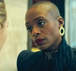 Mulher de cabelo raspado, blazer listrado e brinco dourado, olha intensamente para loira de costas. Cenário interno e sério.