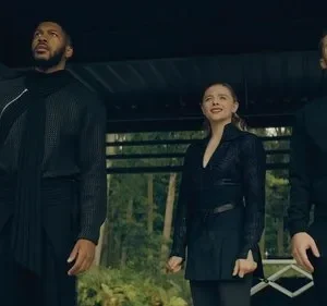 Quatro pessoas olham para cima com expressões sérias. Homem e mulher com roupas escuras e futuristas, outra mulher com casaco xadrez. Ambiente natural.