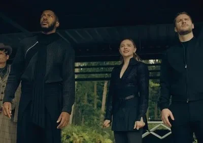 Quatro pessoas olham para cima com expressões sérias. Homem e mulher com roupas escuras e futuristas, outra mulher com casaco xadrez. Ambiente natural.