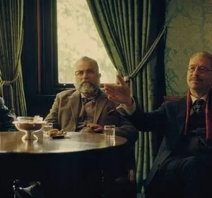 Três homens idosos em trajes vintage sentados ao redor de uma mesa em um ambiente clássico, um deles gesticulando. Clima formal.