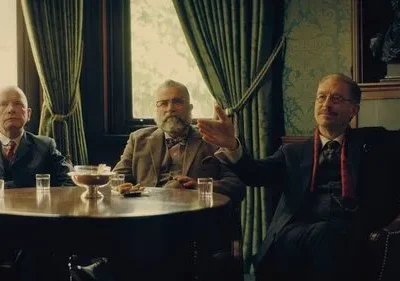 Três homens idosos em trajes vintage sentados ao redor de uma mesa em um ambiente clássico, um deles gesticulando. Clima formal.