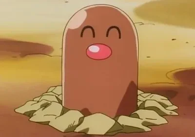 Uma criatura marrom cilíndrica com nariz rosa e olhos fechados, sorrindo, emerge da terra em um cenário rochoso.