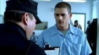 Oficial de uniforme escuro olha para baixo; jovem detento de camisa azul clara o observa em ambiente prisional. Clima tenso.