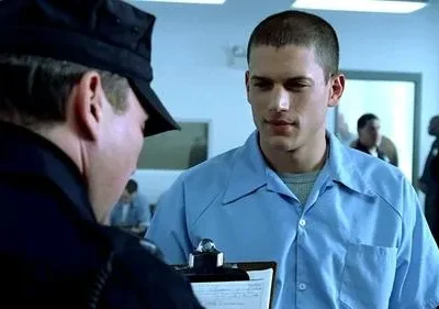 Oficial de uniforme escuro olha para baixo; jovem detento de camisa azul clara o observa em ambiente prisional. Clima tenso.