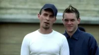 Dois homens em close-up. Um de boné tem expressão tensa. O outro, de jaqueta azul e cabelo espetado, sorri.