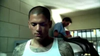Jovem tatuado e sério de regata branca em cela. Ao fundo, outro homem escreve atrás das grades. Ambiente escuro e tenso.