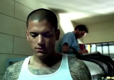 Jovem tatuado e sério de regata branca em cela. Ao fundo, outro homem escreve atrás das grades. Ambiente escuro e tenso.