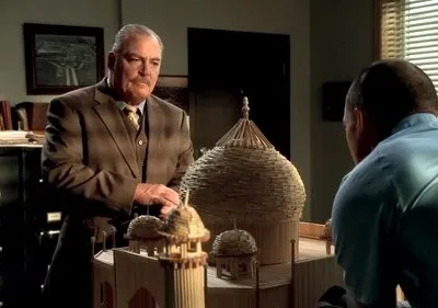 Dois homens em um escritório examinam um grande modelo de edifício feito de palitos de artesanato. Um clima concentrado e sério.