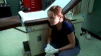 Mulher agachada no chão de um consultório médico, com expressão angustiada e luvas brancas. Um ambiente clínico e tenso.