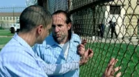 Dois homens em uniformes azuis conversam perto de uma cerca de arame. Estão em um pátio de prisão, com edifícios e guardas ao fundo.