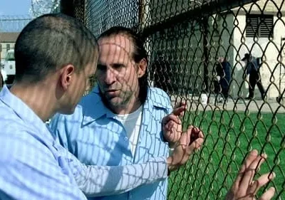 Dois homens em uniformes azuis conversam perto de uma cerca de arame. Estão em um pátio de prisão, com edifícios e guardas ao fundo.