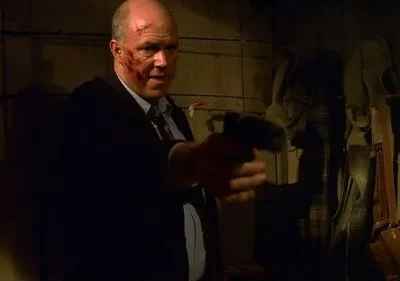 Homem careca e ferido, de terno escuro, aponta uma pistola num cômodo rústico e escuro, com objetos pendurados. Clima de perigo.