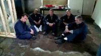 Cinco homens em uniformes azuis sentados num chão úmido e sujo, num quarto escuro e desgastado. Seus rostos expressam seriedade.