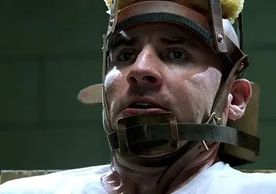 Close-up de homem assustado, usando aparelho de contenção na cabeça. Fundo institucional, clima de aflição e tensão.
