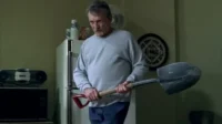 Um homem idoso e sério, vestindo cinza, segura uma pá, parado em uma cozinha simples com geladeira. O clima é tenso e ameaçador.
