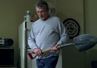 Um homem idoso e sério, vestindo cinza, segura uma pá, parado em uma cozinha simples com geladeira. O clima é tenso e ameaçador.